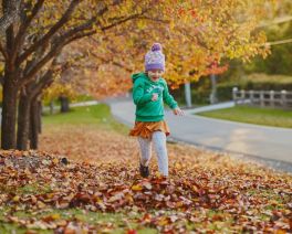 A girl wearing a bright green jumper and beanie running throughout a ground covered in fallen autumn leaves.
